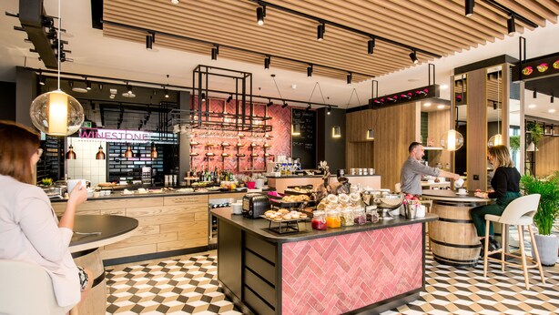 Un café moderne avec un sol à carreaux, un bar central dont le comptoir présente un motif en chevron rose, et une enseigne indiquant la présence de vins, bien visible.