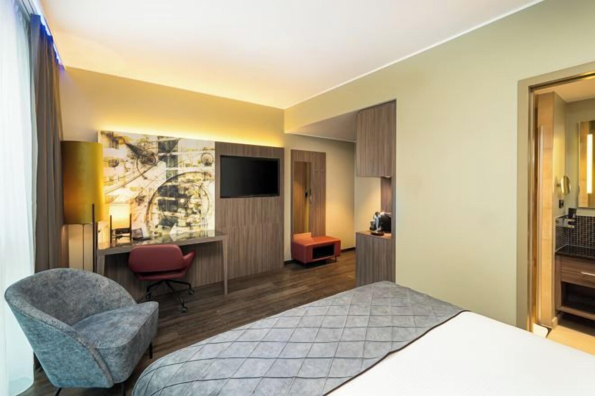 A hotel room with a bed, a desk with a chair, a TV, and a small seating area.