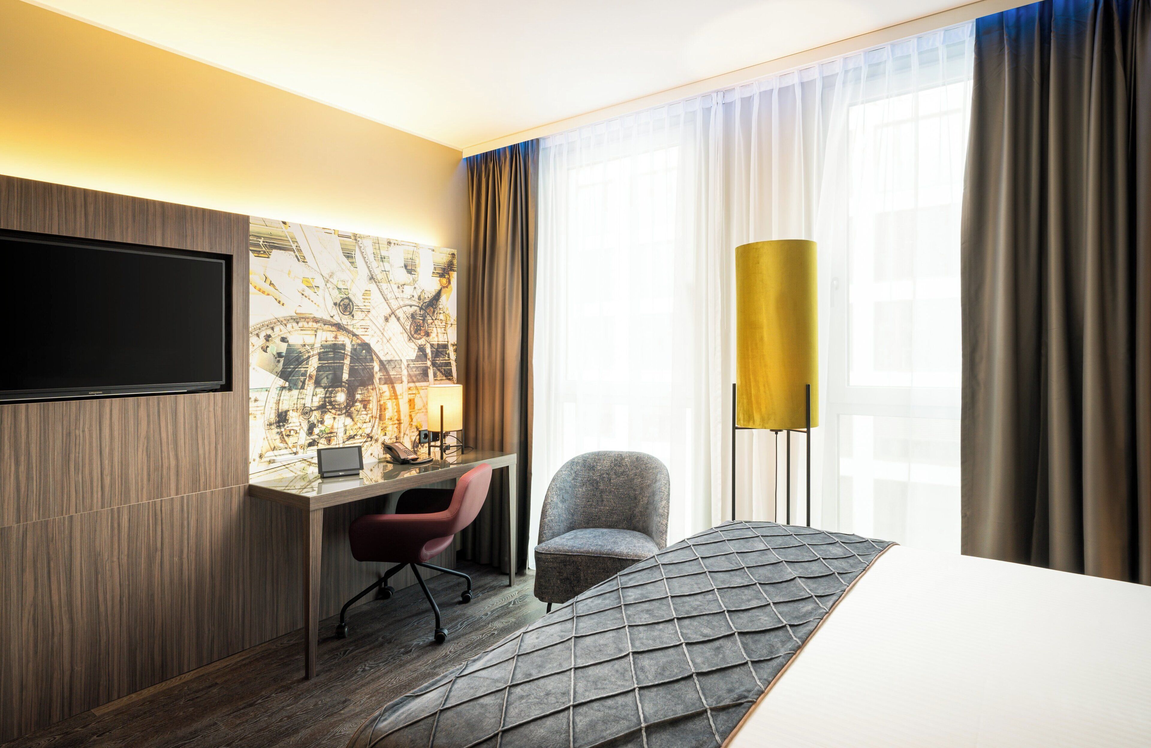 Una habitación de hotel con un televisor de pantalla plana en la pared, un escritorio con una computadora portátil, un sillón cómodo, un ventanal con cortinas y una alfombra estampada.
