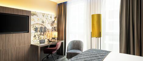 Una habitación de hotel con un televisor de pantalla plana en la pared, un escritorio con una computadora portátil, un sillón cómodo, un ventanal con cortinas y una alfombra estampada.