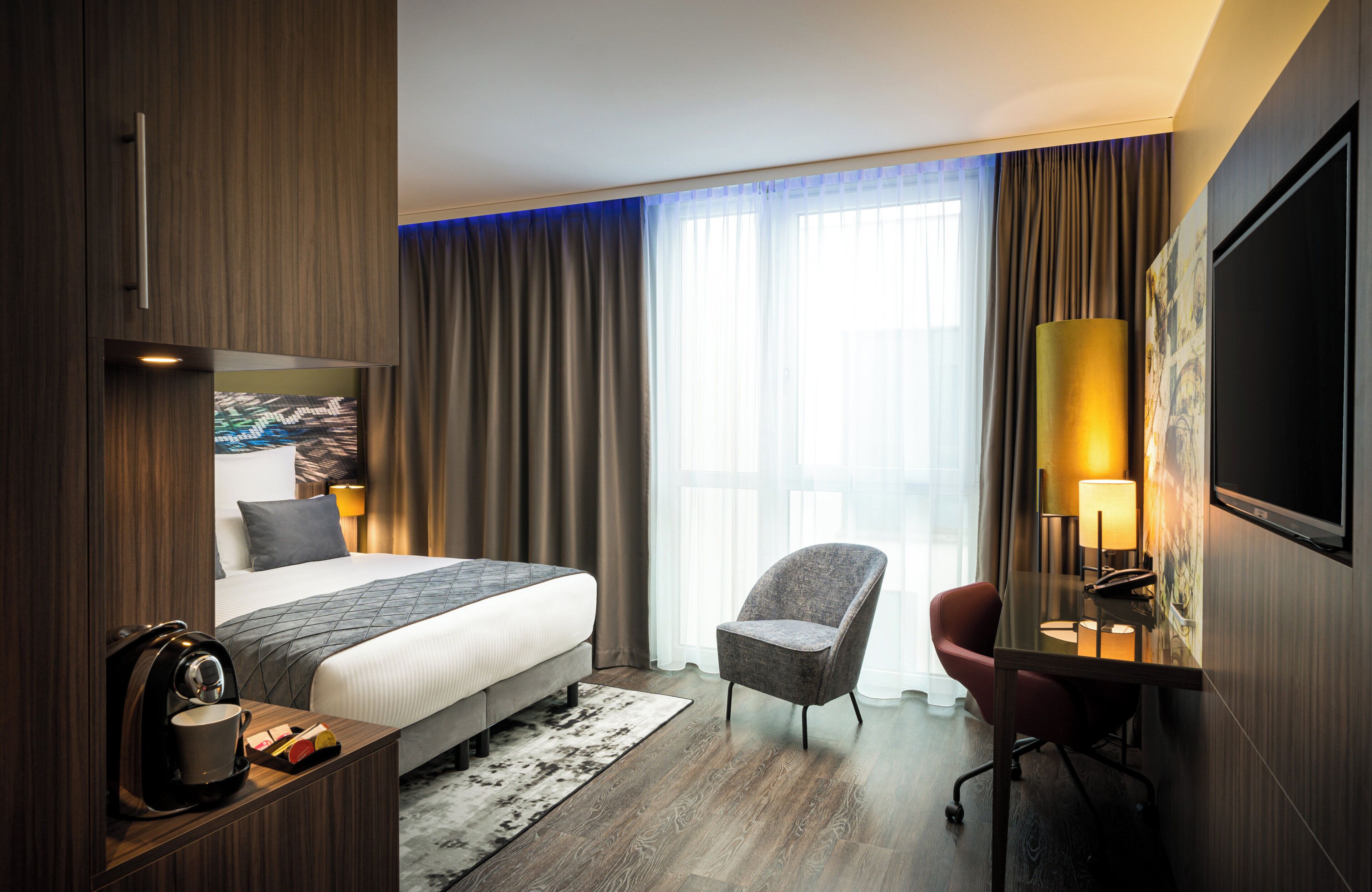 A hotel room with a bed, a desk with a lamp, a chair, and a TV mounted on the wall.