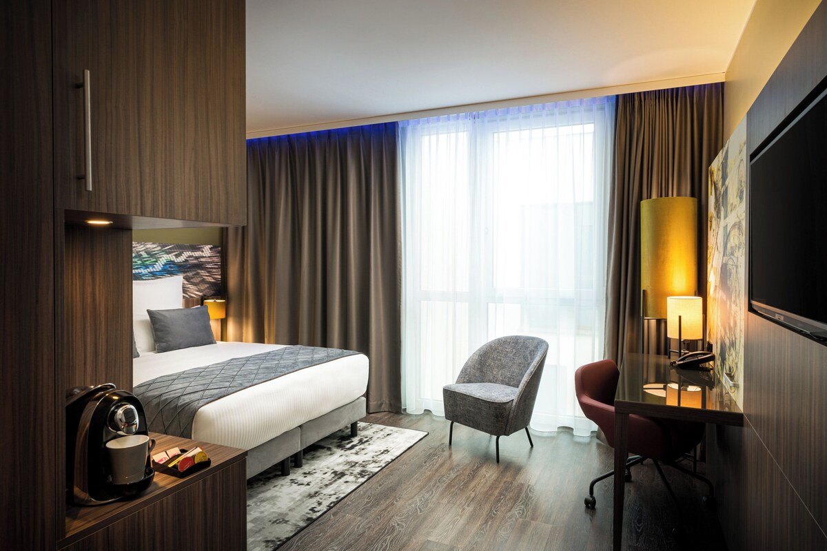 A hotel room with a bed, a desk with a lamp, a chair, and a TV mounted on the wall.