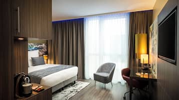 A hotel room with a bed, a desk with a lamp, a chair, and a TV mounted on the wall.