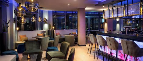 A modern bar area with seating and a bar counter, featuring pendant lighting and a view of the cityscape.