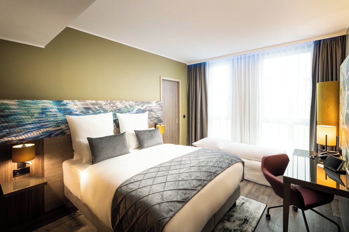 A hotel room with a large bed, a desk with a chair, a lamp, and a view of the outdoors.