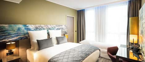 A hotel room with a large bed, a desk with a chair, a lamp, and a view of the outdoors.