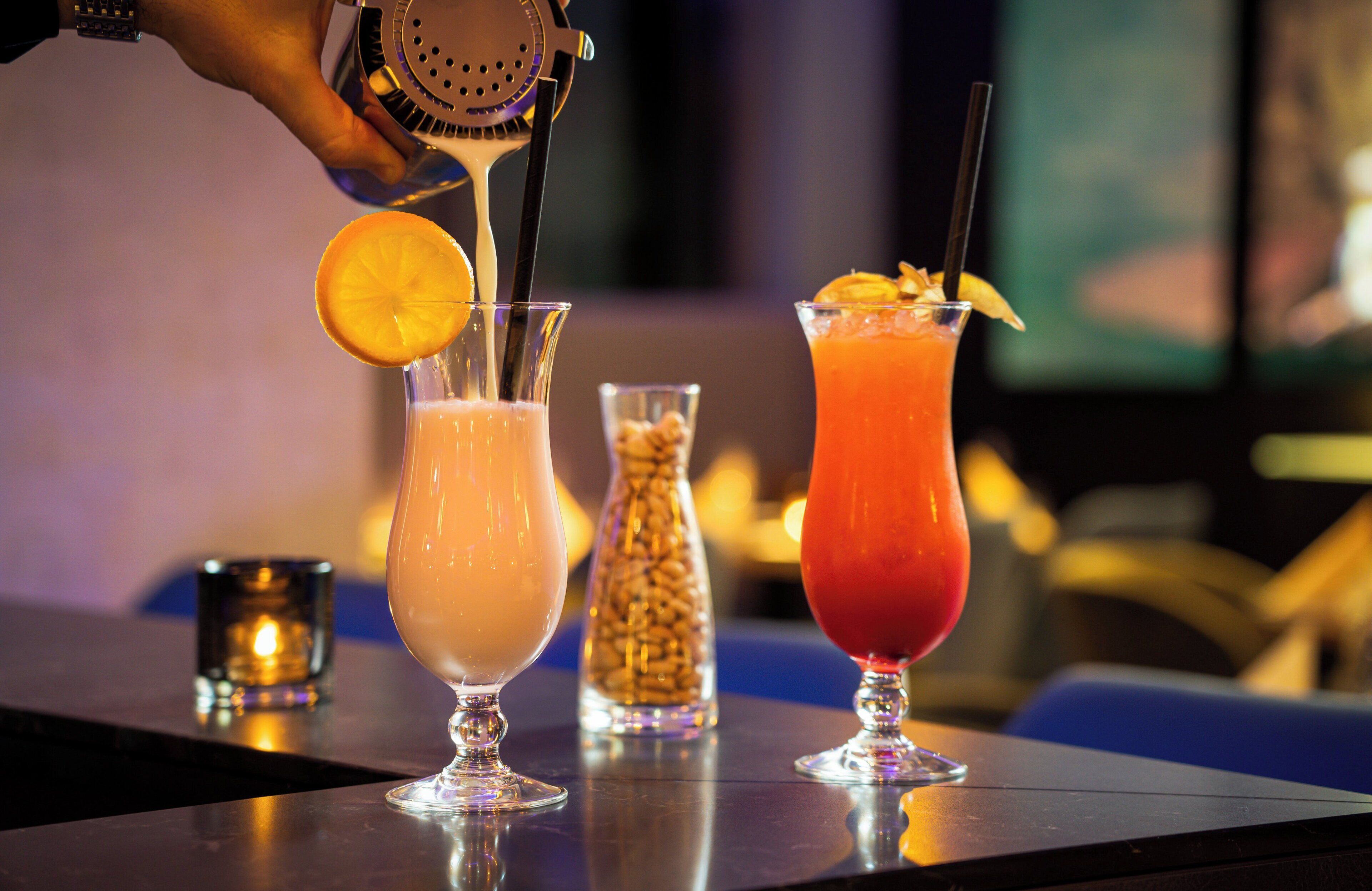 Three cocktails on a bar counter with a hand pouring a drink into one of them.
