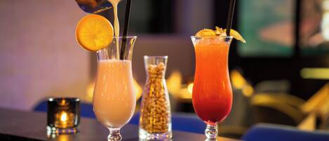 Three cocktails on a bar counter with a hand pouring a drink into one of them.