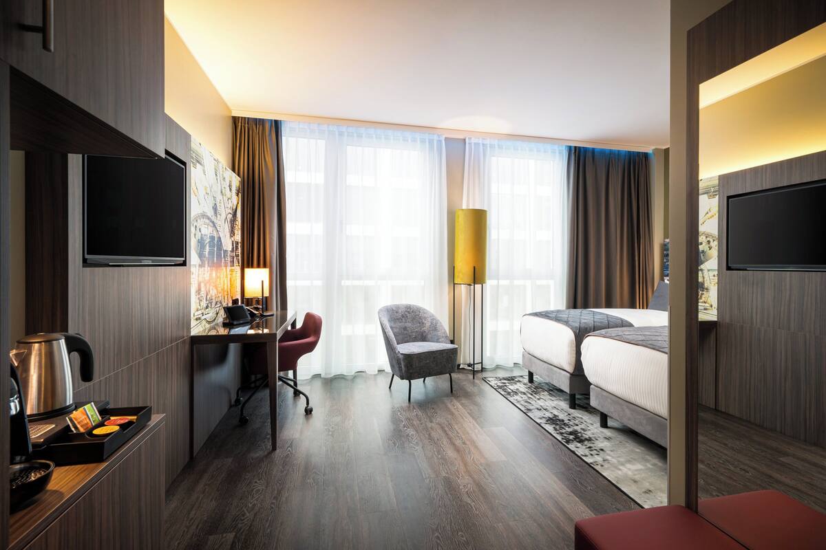 Et hotelværelse med en seng, et skrivebord med stol, et fjernsyn, en lampe og et stort vindue med gardiner.