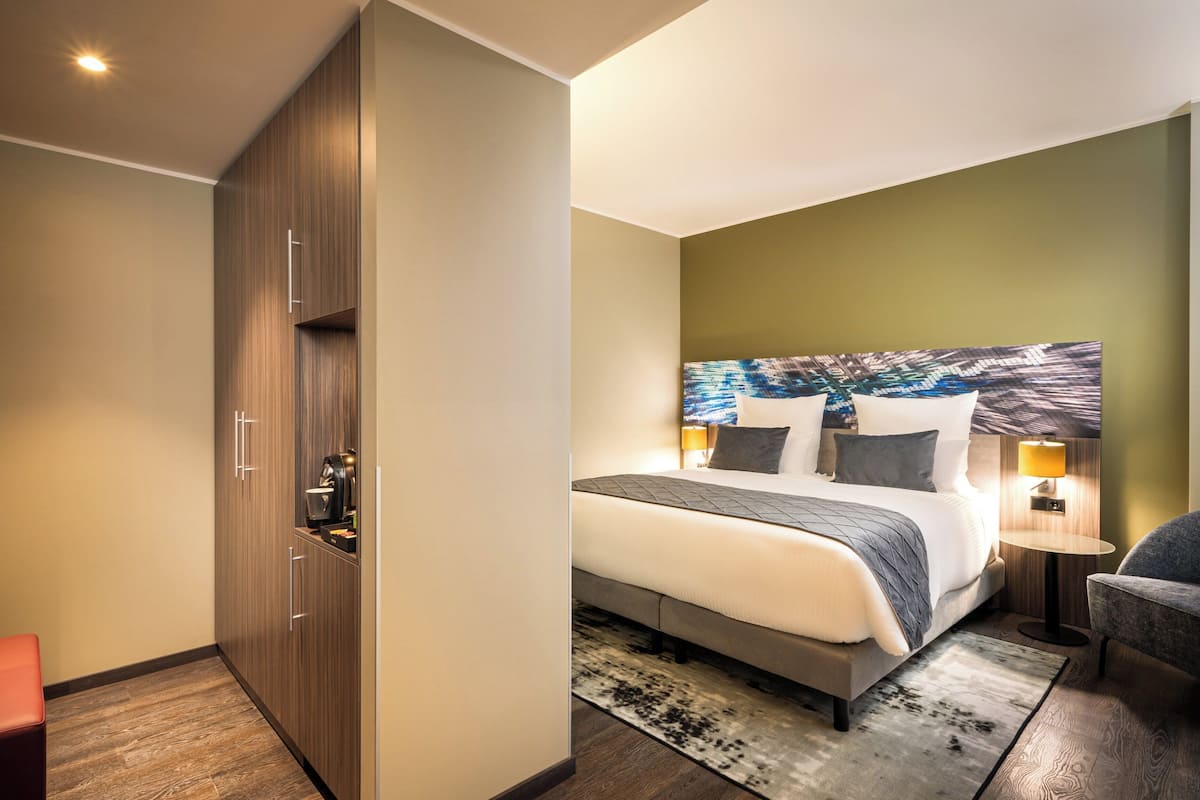 Et moderne hotelværelse med en stor seng, et natbord med lampe, et siddeområde med en stol og et garderobeskab.