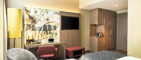 A hotel room with a bed, a desk with a computer, a TV, and a seating area with chairs.