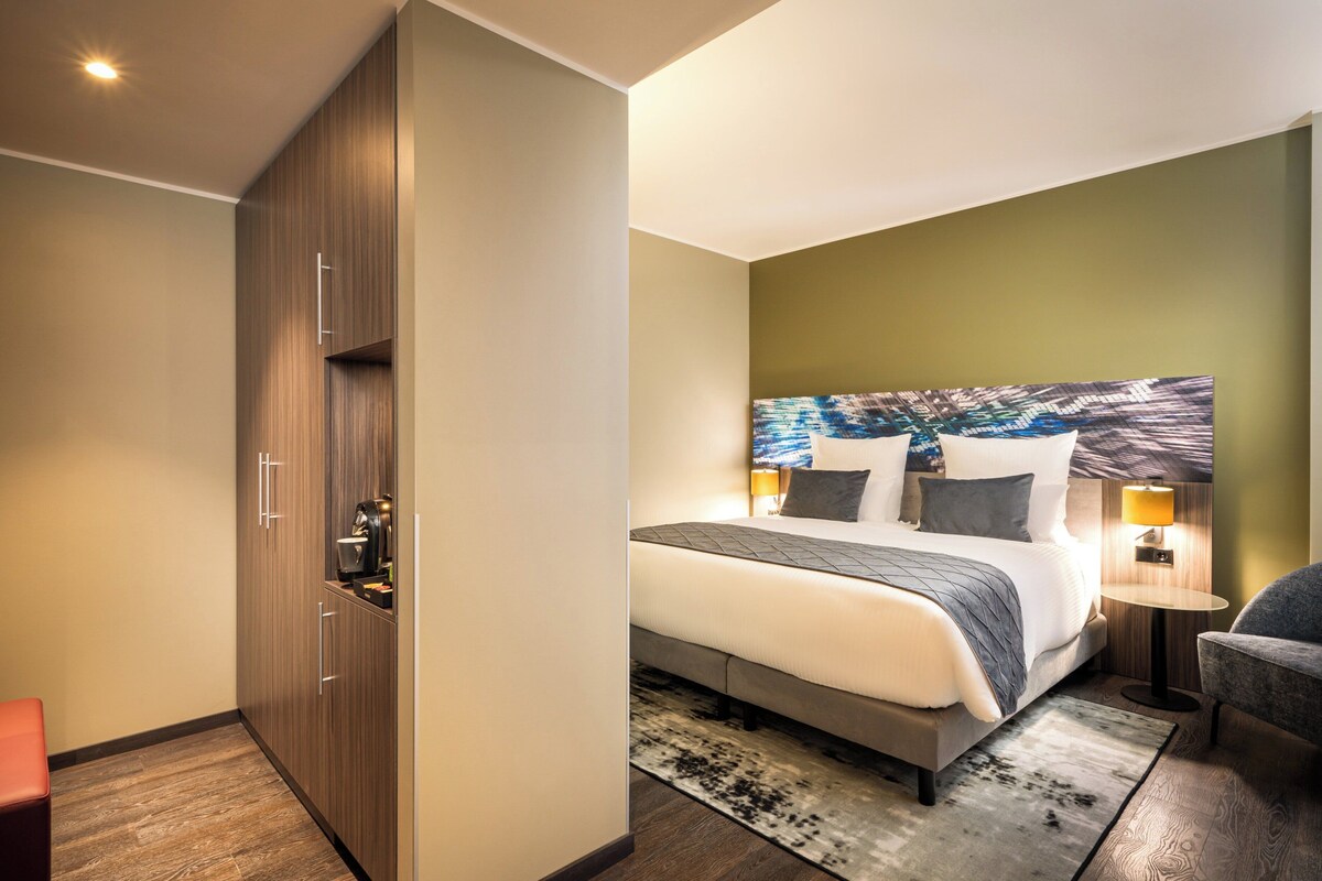 A modern hotel room with a large bed, a nightstand with a lamp, a seating area with a chair, and a wardrobe.