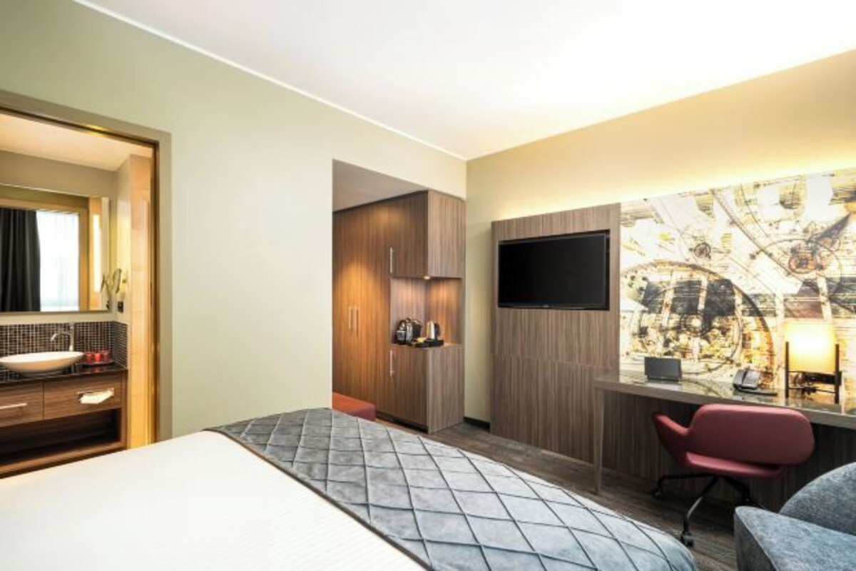 A hotel room with a bed, a desk with a chair, a TV, a bathroom with a sink and mirror, and a wardrobe.