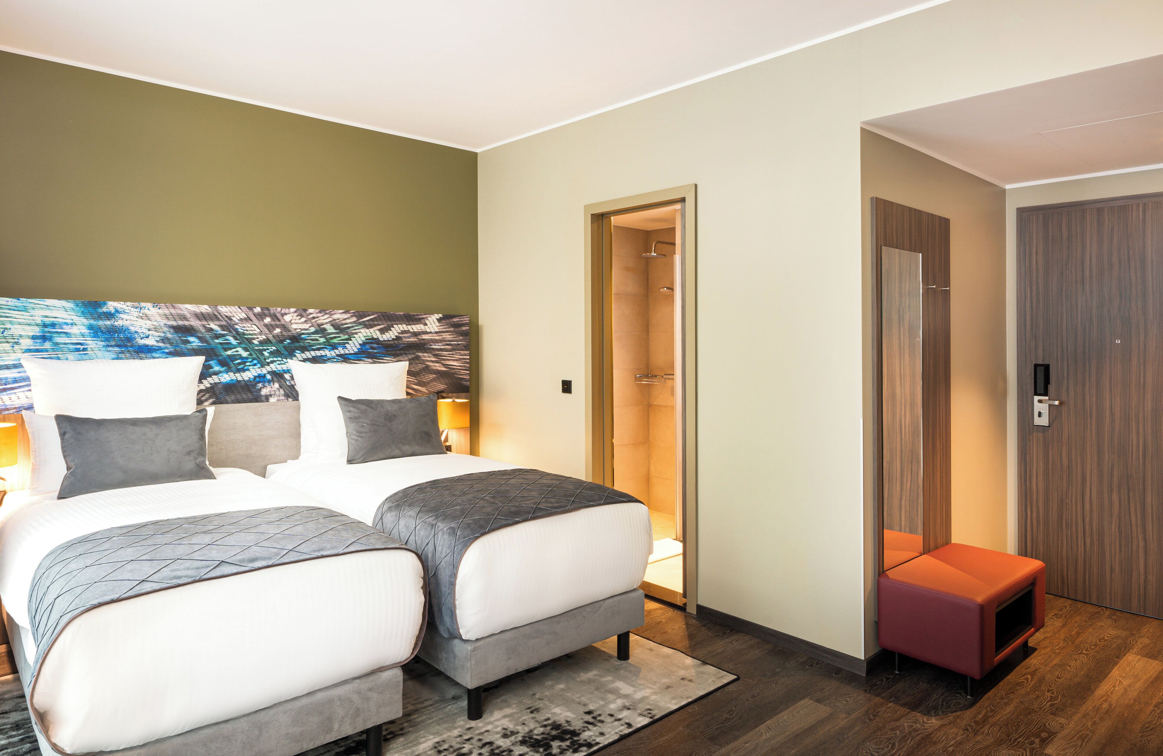 Habitación de hotel con dos camas, un cuadro abstracto grande en la pared, una puerta de madera y un pequeño banco rojo.