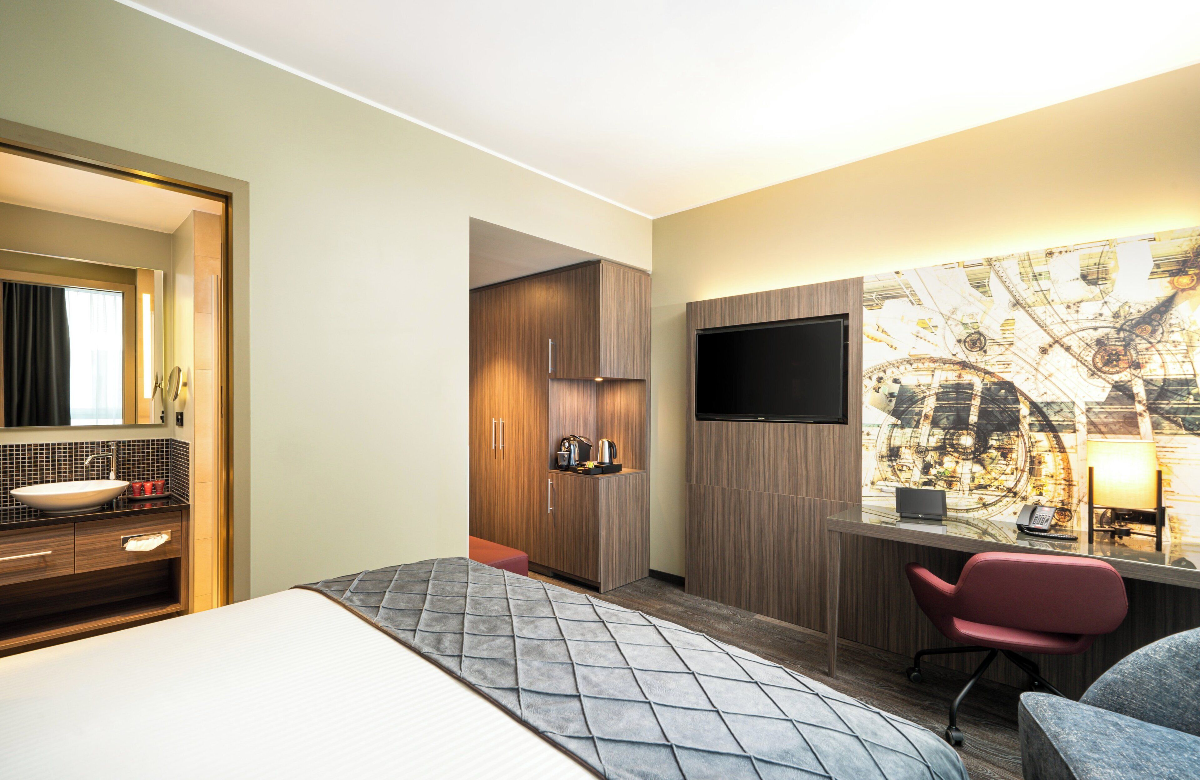Habitación de hotel con cama, escritorio con silla, televisor, baño con lavamanos y espejo.
