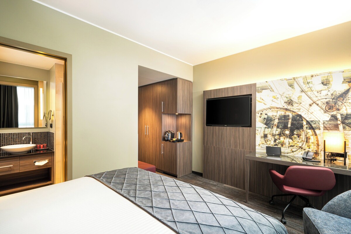 A hotel room with a bed, a desk with a chair, a TV, a bathroom with a sink, and a mirror.
