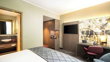 Habitación de hotel con cama, escritorio con silla, televisor, baño con lavamanos y espejo.