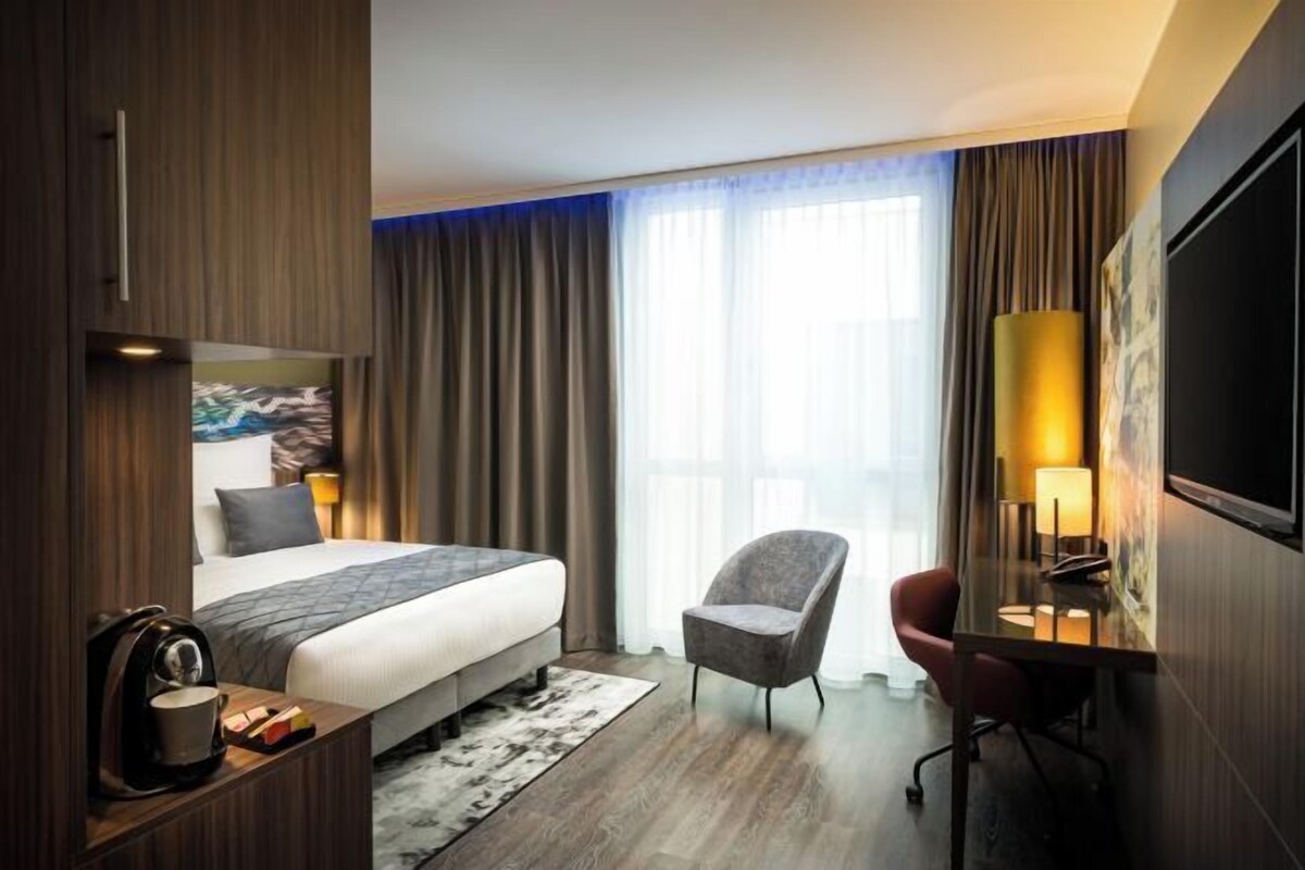 A hotel room with a bed, a desk, a chair, a TV, and a window with curtains.