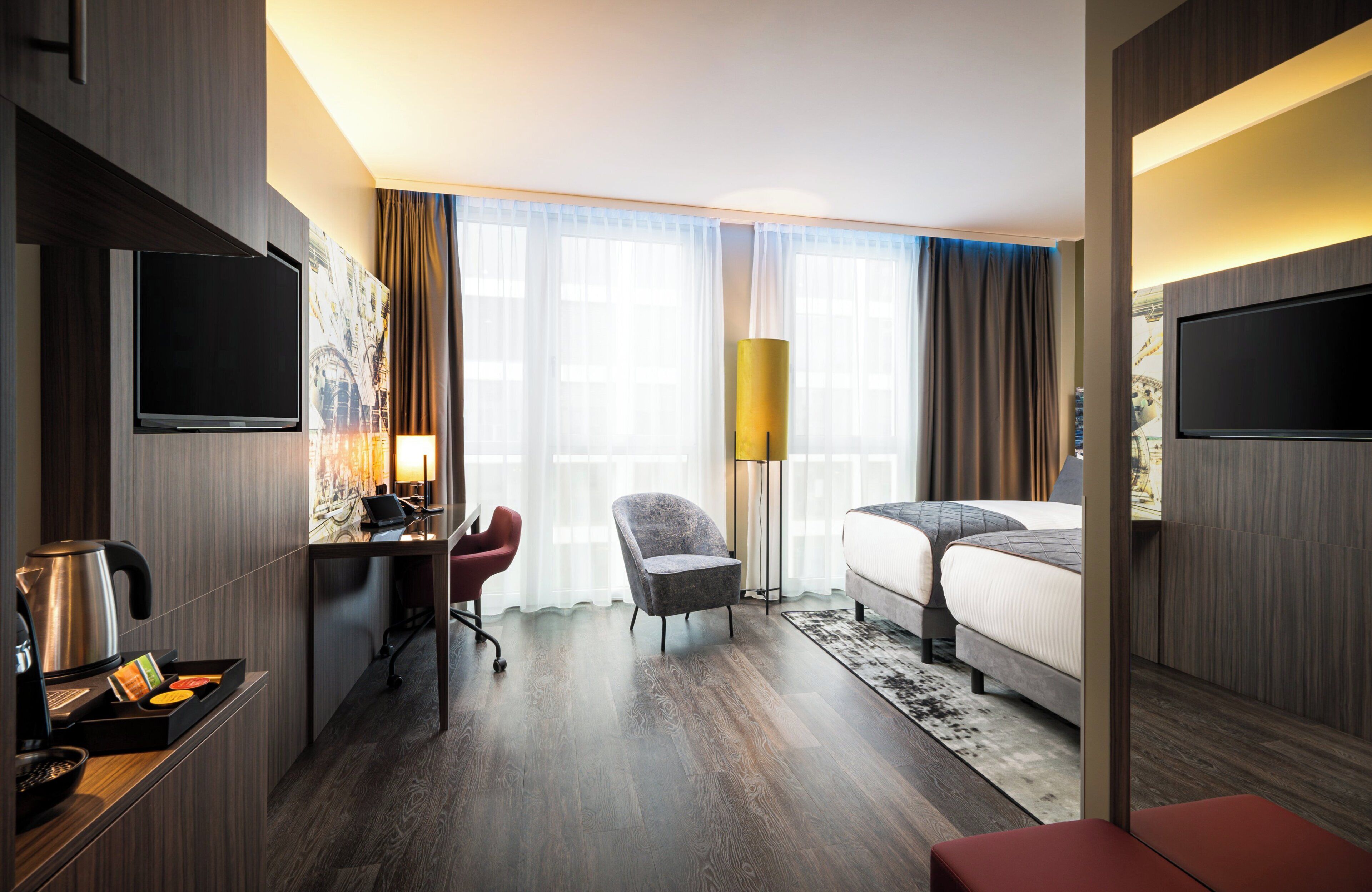 A hotel room with a bed, a desk with a chair, a TV, a lamp, and a large window with curtains.