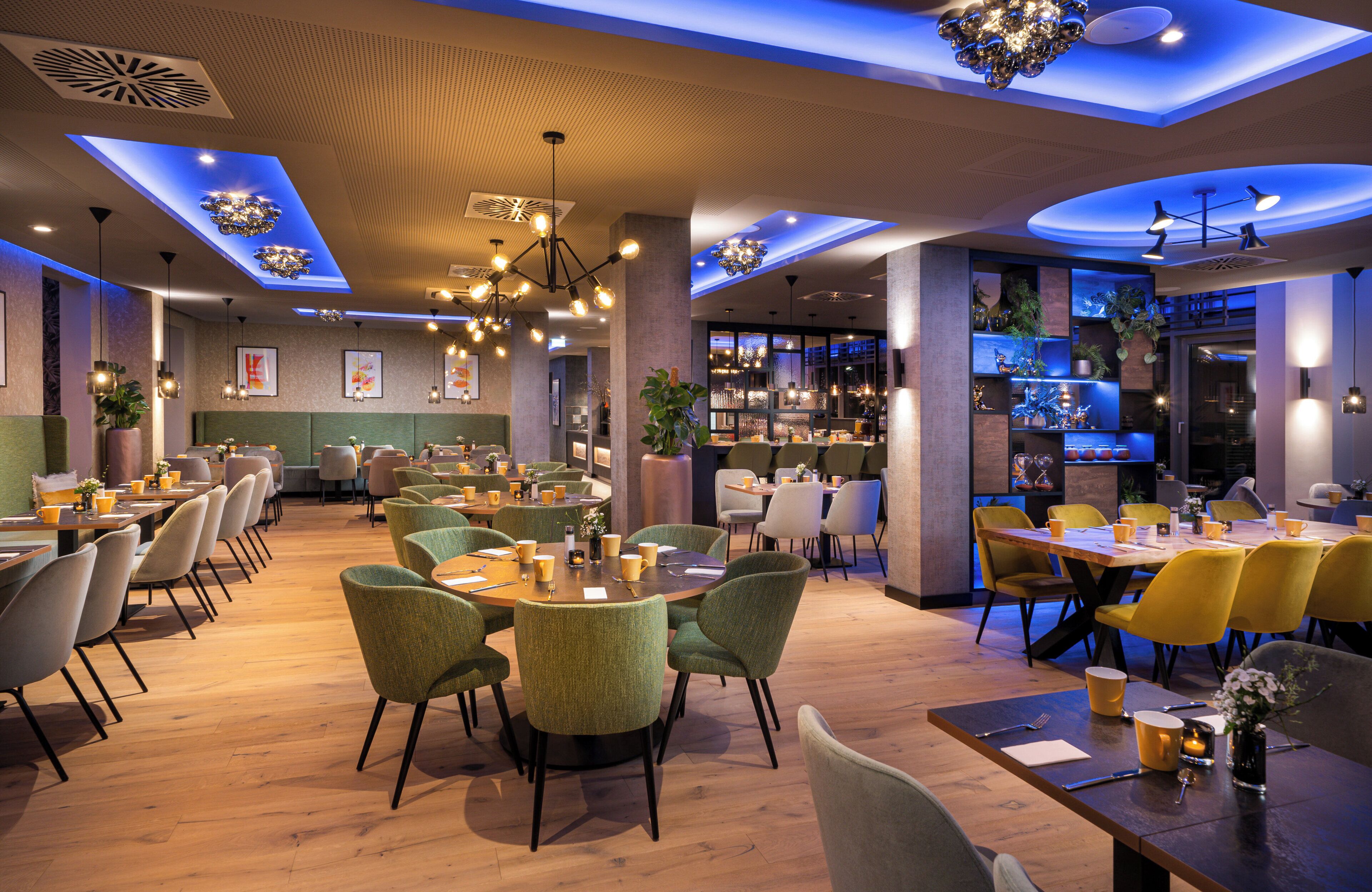 A modern restaurant with round tables, green chairs, and a bar area with shelves of bottles.