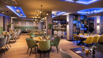 A modern restaurant with round tables, green chairs, and a bar area with shelves of bottles.