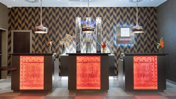 Un área de recepción con tres mostradores, cada uno con un panel iluminado en rojo y una lámpara colgante. Se ve un letrero que dice 'Leonardo' en la pared.