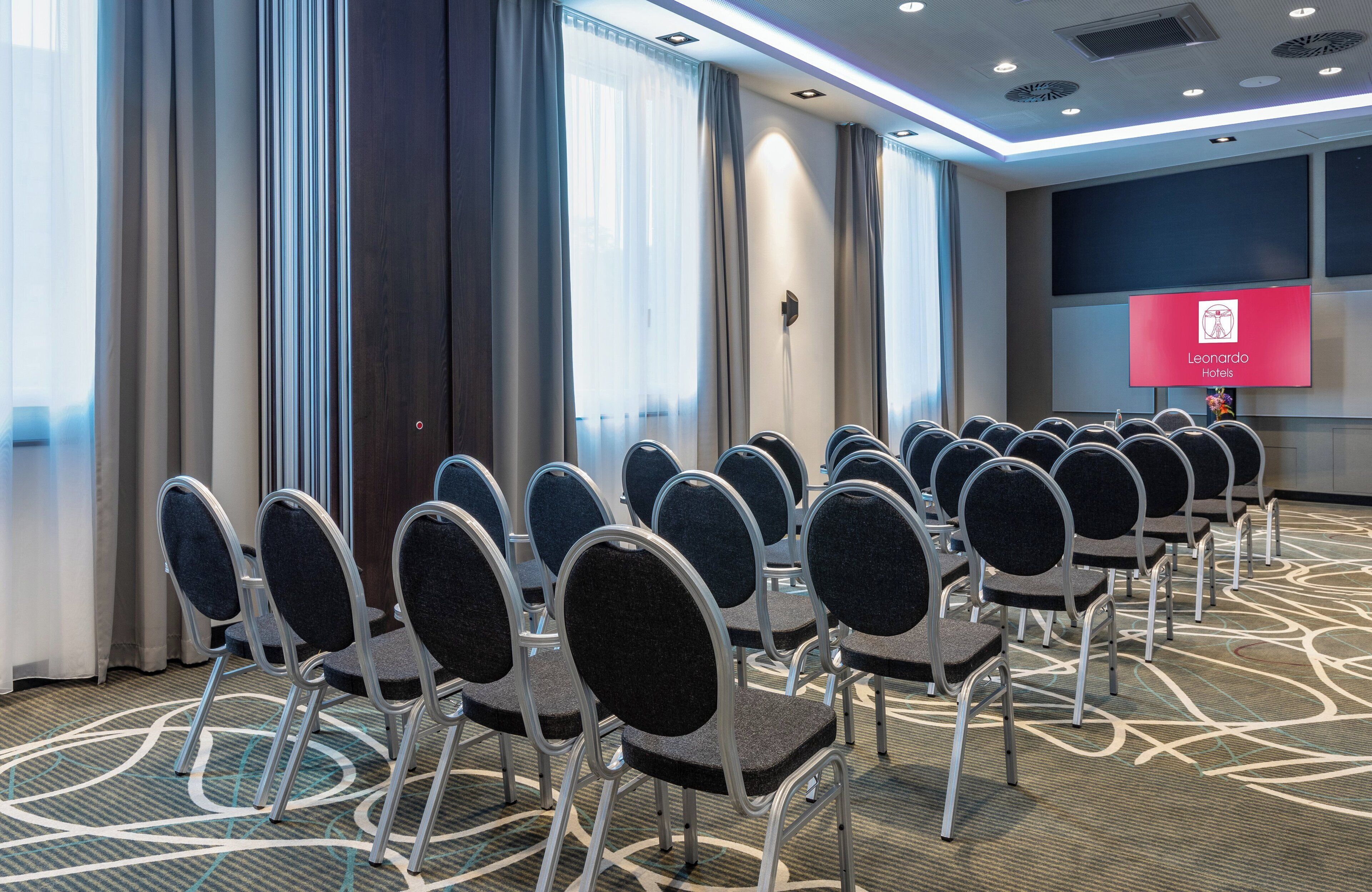 Une salle de conférence avec des rangées de chaises, un grand écran affichant le nom de l’hôtel et un sol recouvert de moquette.