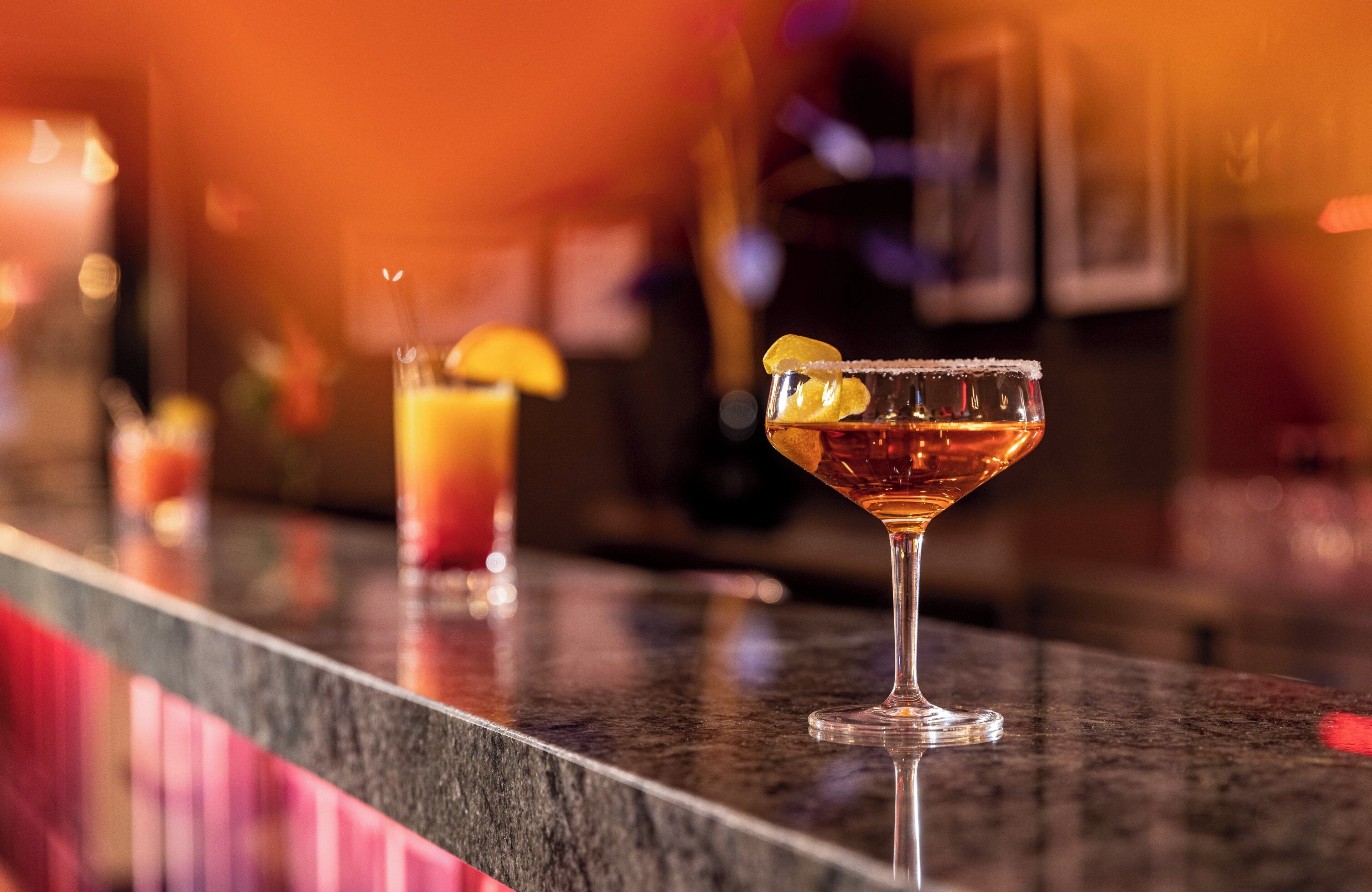 A bar counter with two cocktails, one with a lemon garnish and the other with a lime garnish.