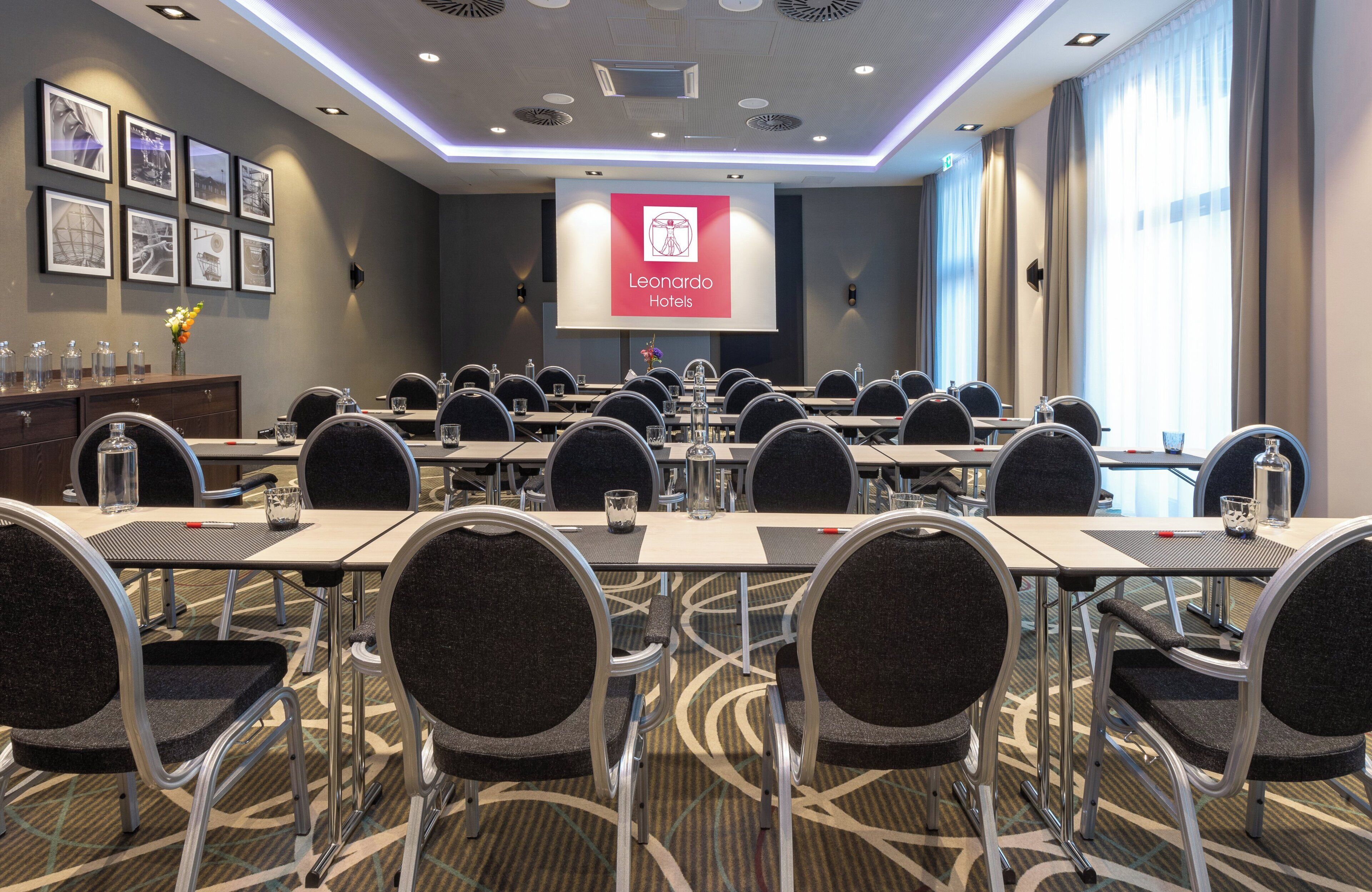 A conference room with tables and chairs, a projector screen displaying the logo of Leonardo Hotels, and framed pictures on the wall.
