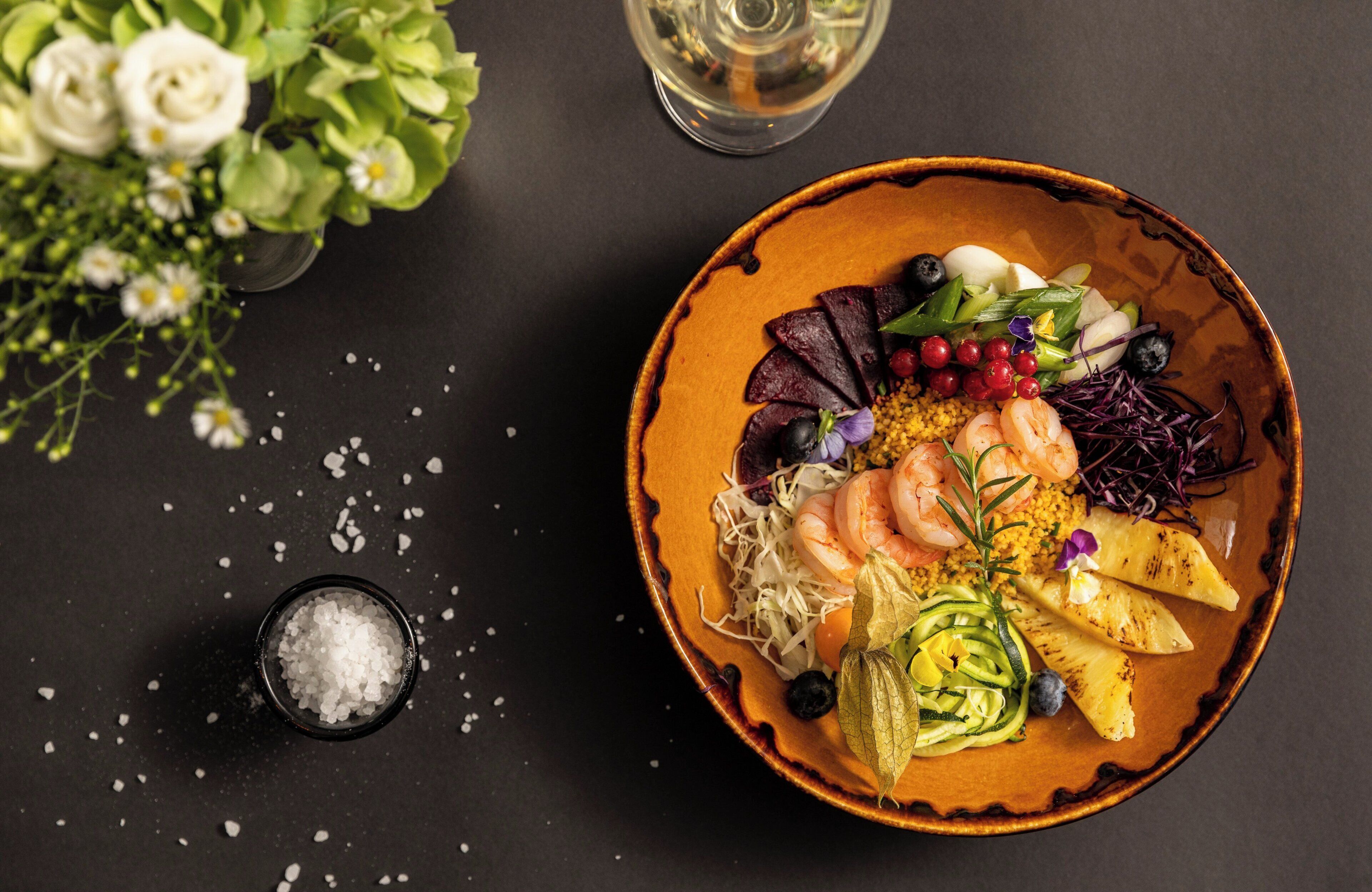 A bowl of food with shrimp, vegetables, and a small bowl of salt on a dark surface.