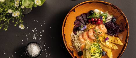 A bowl of food with shrimp, vegetables, and a small bowl of salt on a dark surface.