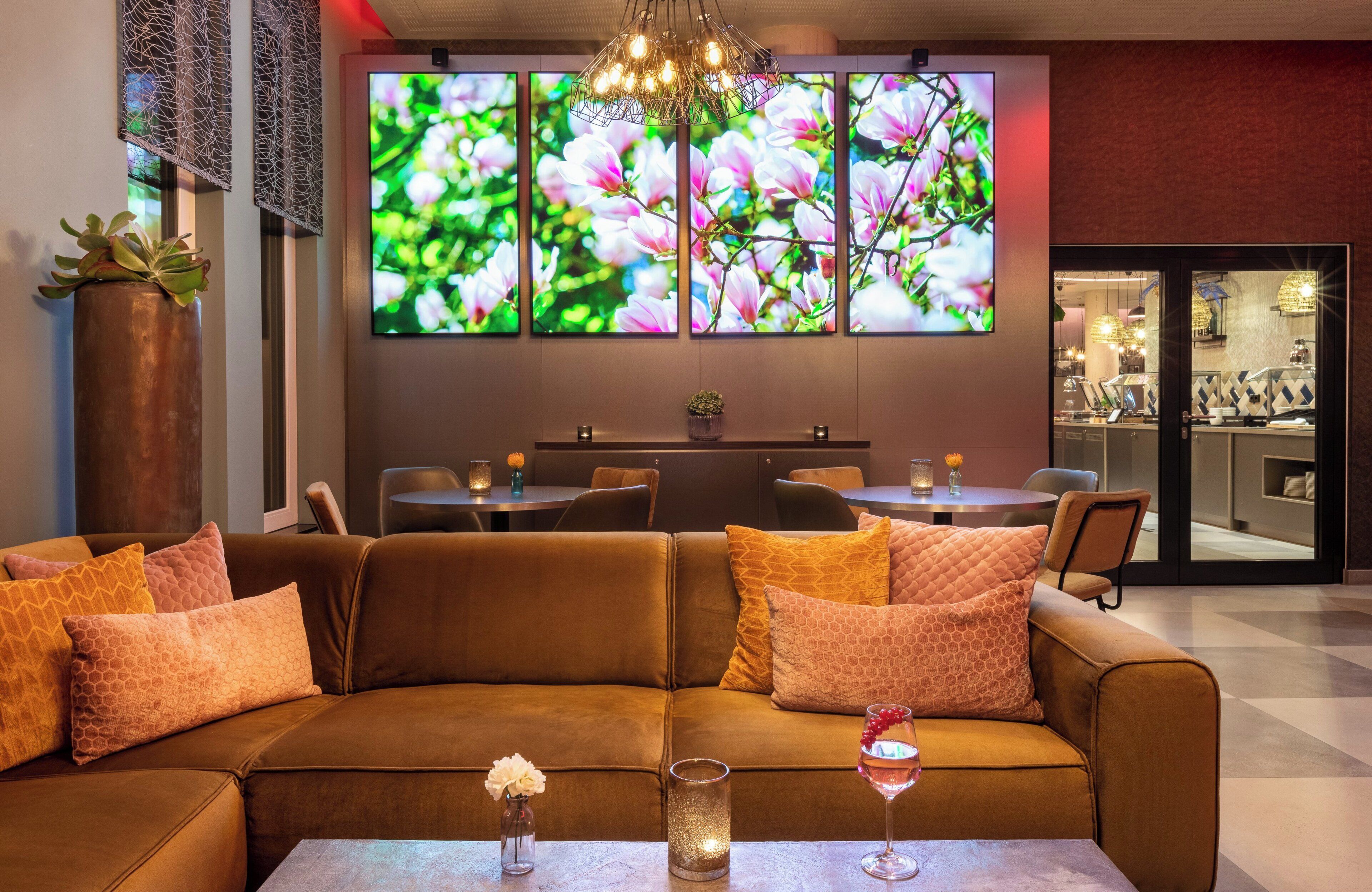 A modern lounge area with a brown leather sofa, multiple cushions, a glass table with a martini glass, and a large floral TV screen.