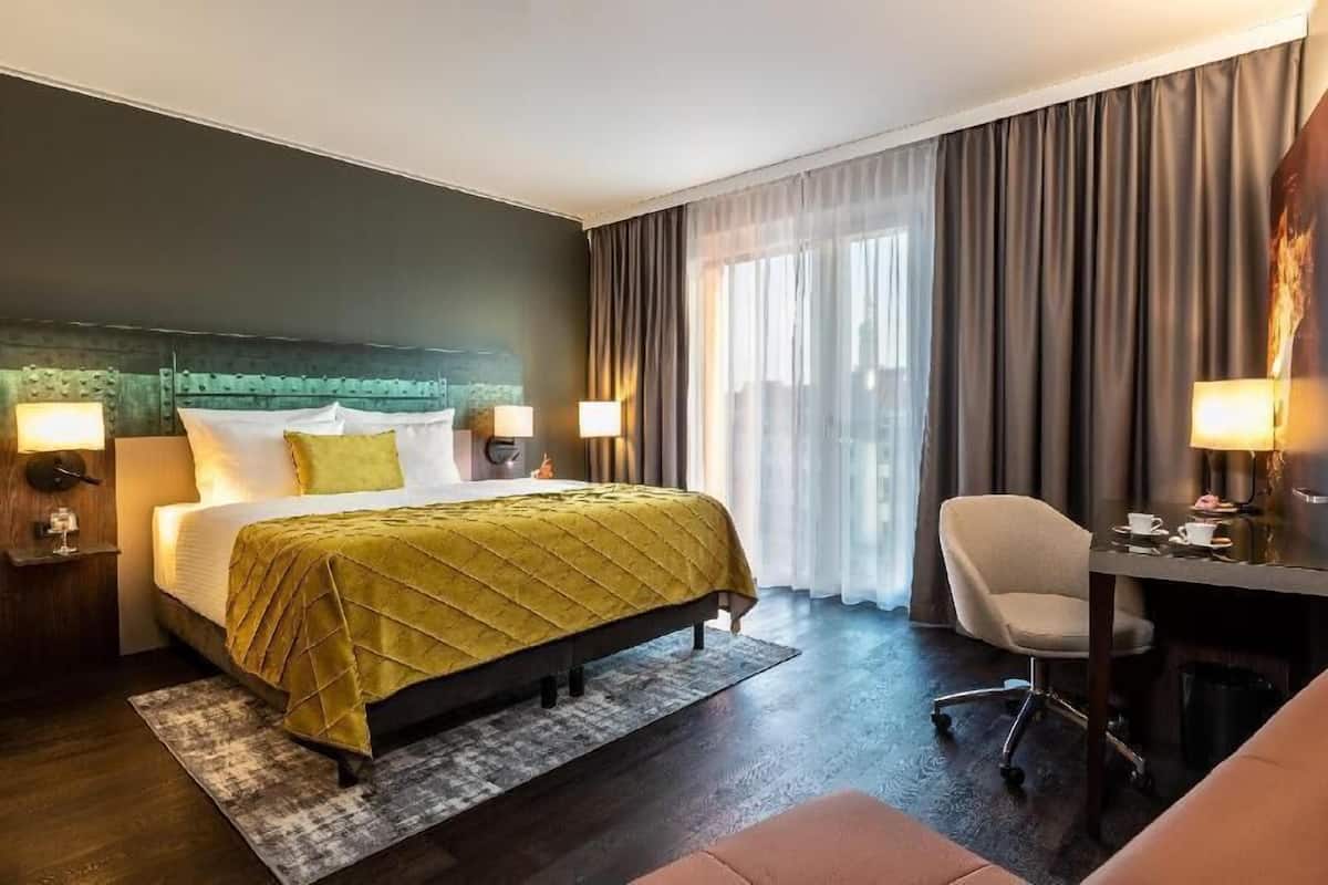 A hotel room with a bed, bedside lamps, a desk with a chair, and a large window with curtains.