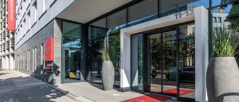 A modern hotel entrance with glass doors, a red carpet, and a large potted plant.