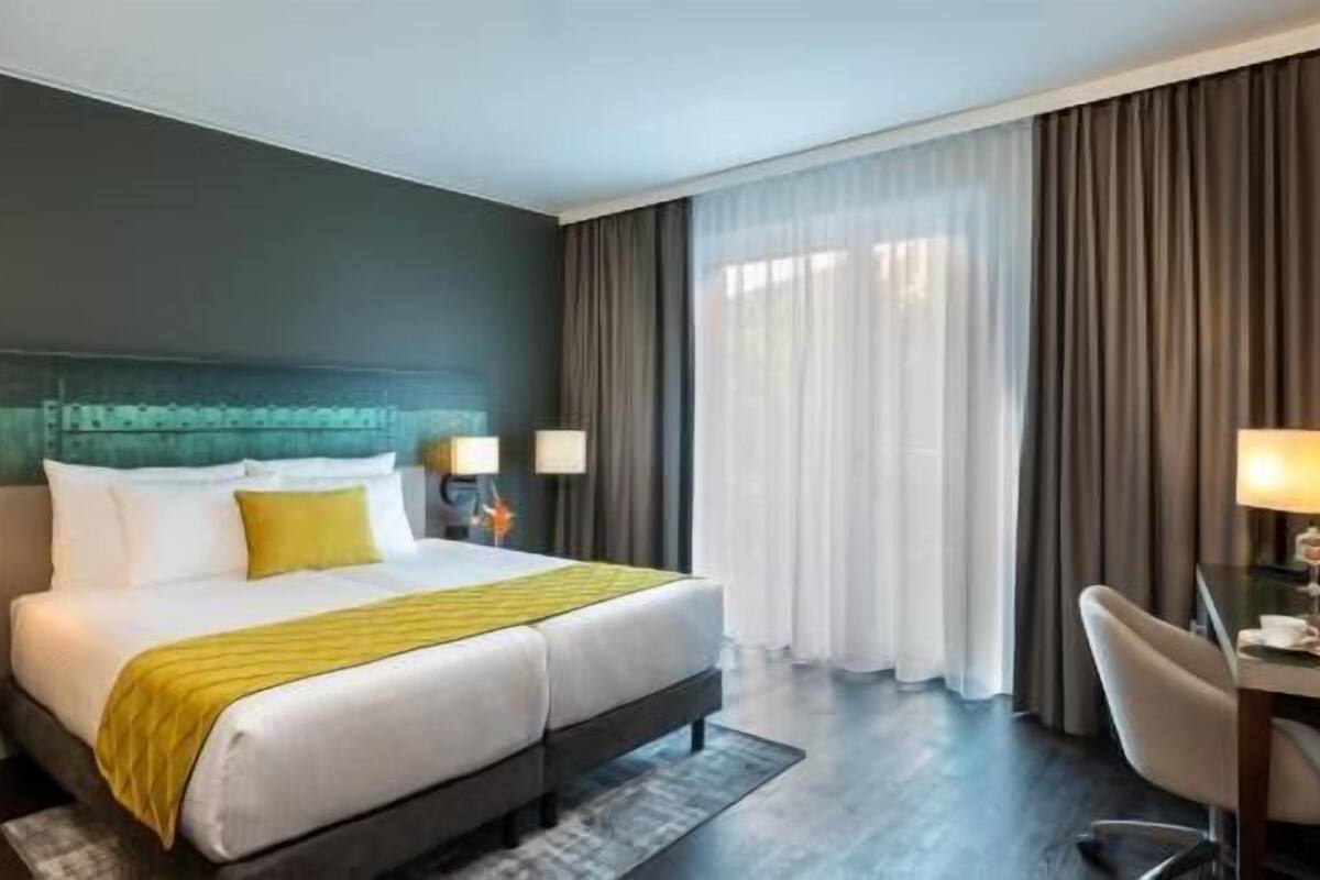 A hotel room with a large bed, two bedside tables with lamps, a desk with a chair, and a view of the outdoors through sheer curtains.