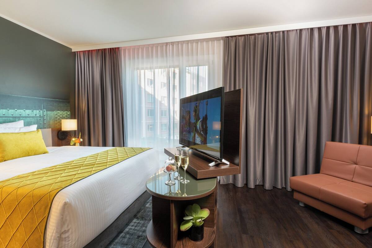 Una habitación de hotel con cama, televisor, una mesita con vasos, sofá y cortinas.
