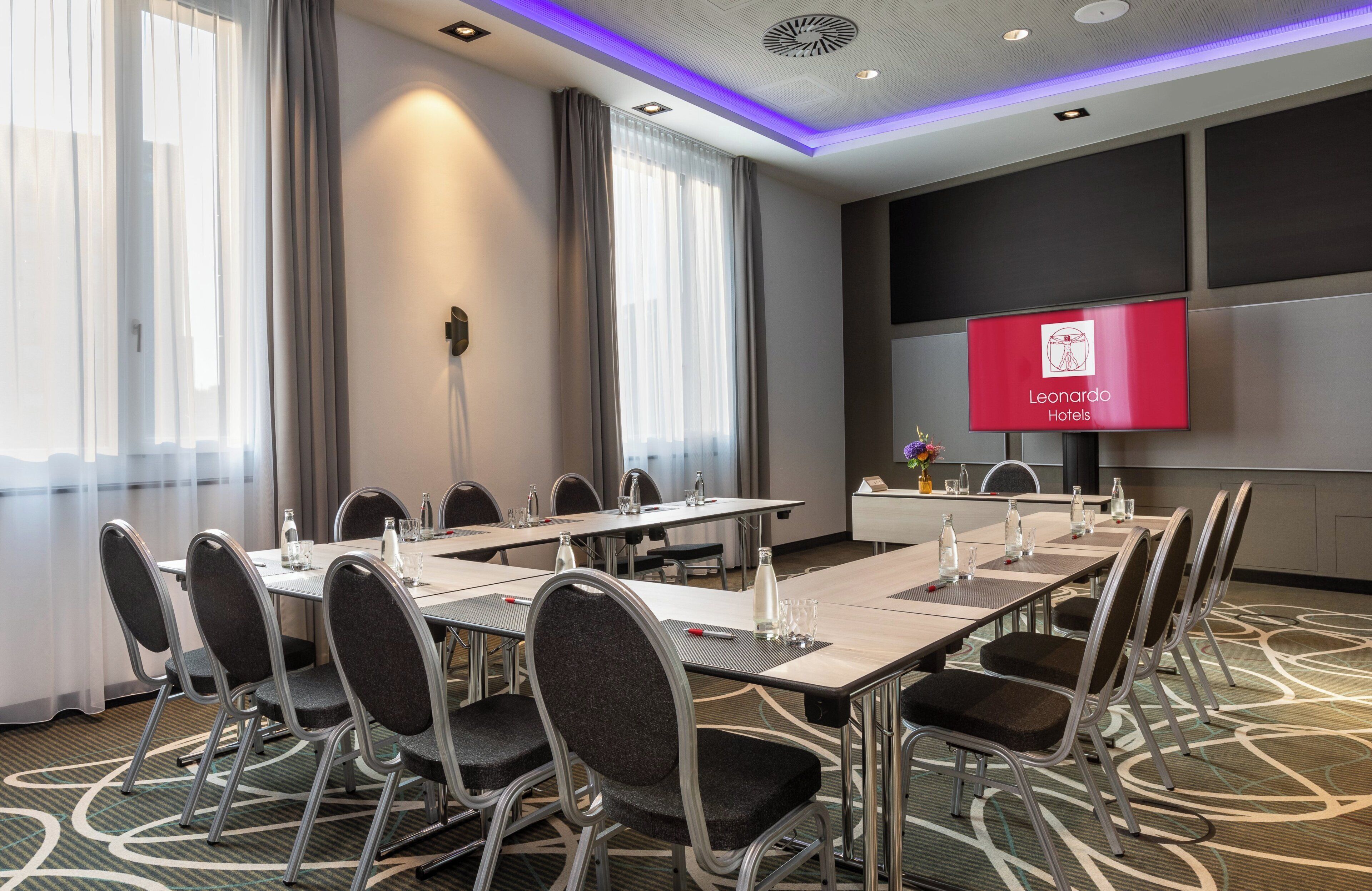 A conference room with a long table, chairs, a flat-screen TV displaying the hotel name, and a large window with curtains.