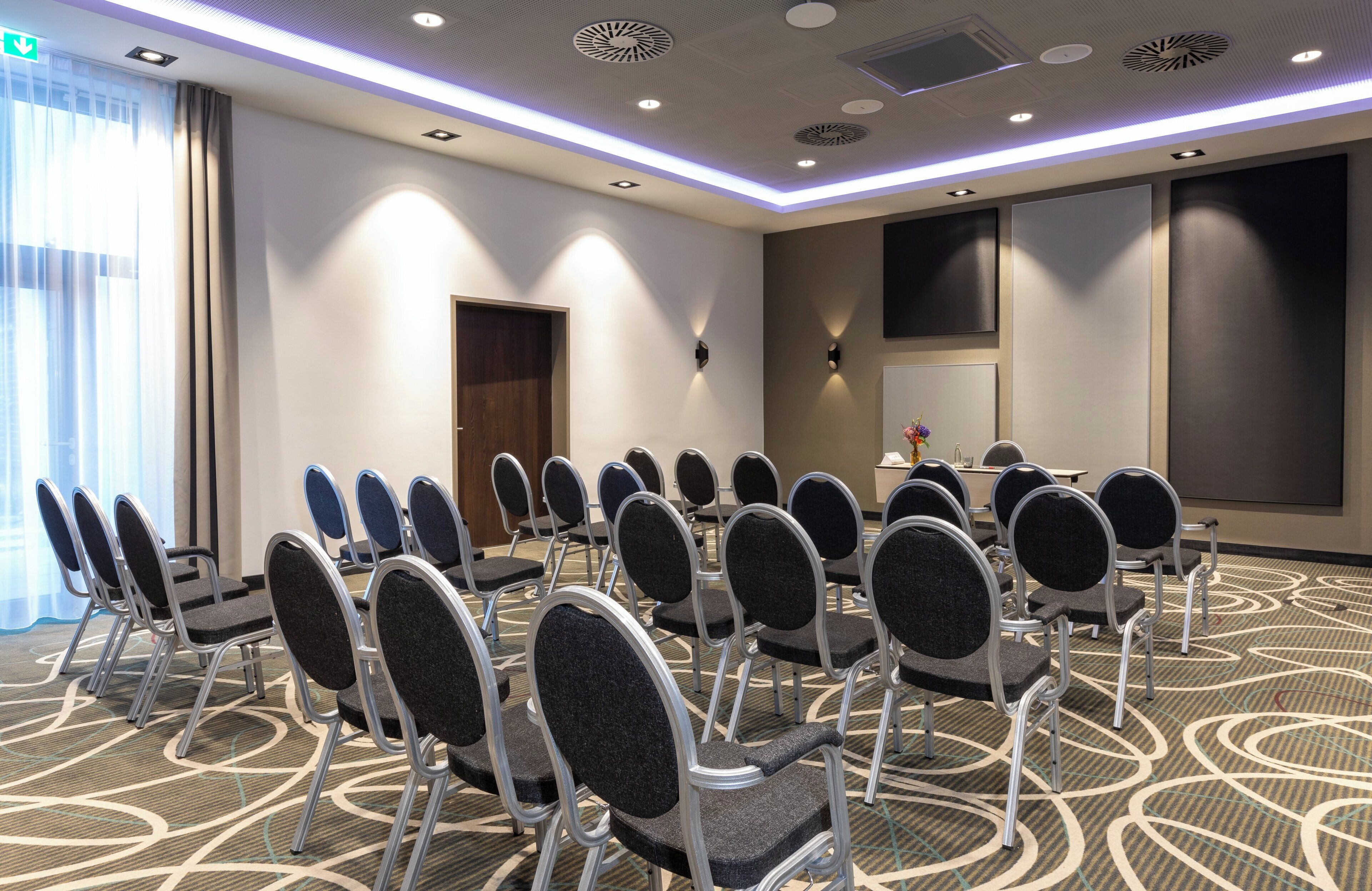 Un salón de conferencias con filas de sillas, una pantalla grande y una puerta.
