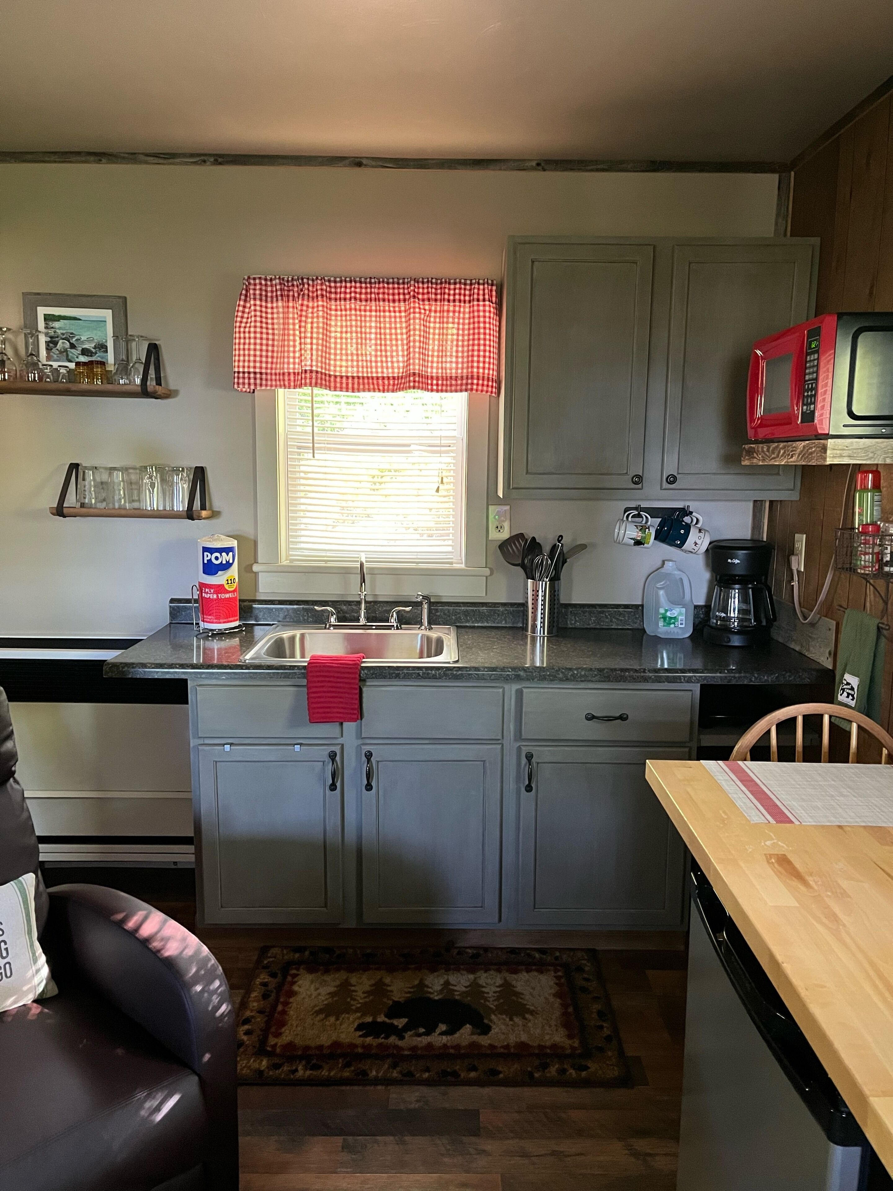 A compact kitchen with a sink, a microwave, and a coffee maker. There is a window with blinds, a red checkered valance, and a wooden chair.