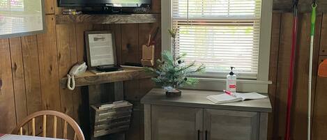 A rustic room with a wooden wall, a mounted television, a small cabinet, a window with blinds, and a broom leaning against the wall.