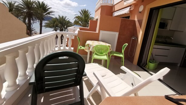 Un balcon avec des chaises blanches et une table, un coin cuisine et, au fond, des palmiers.