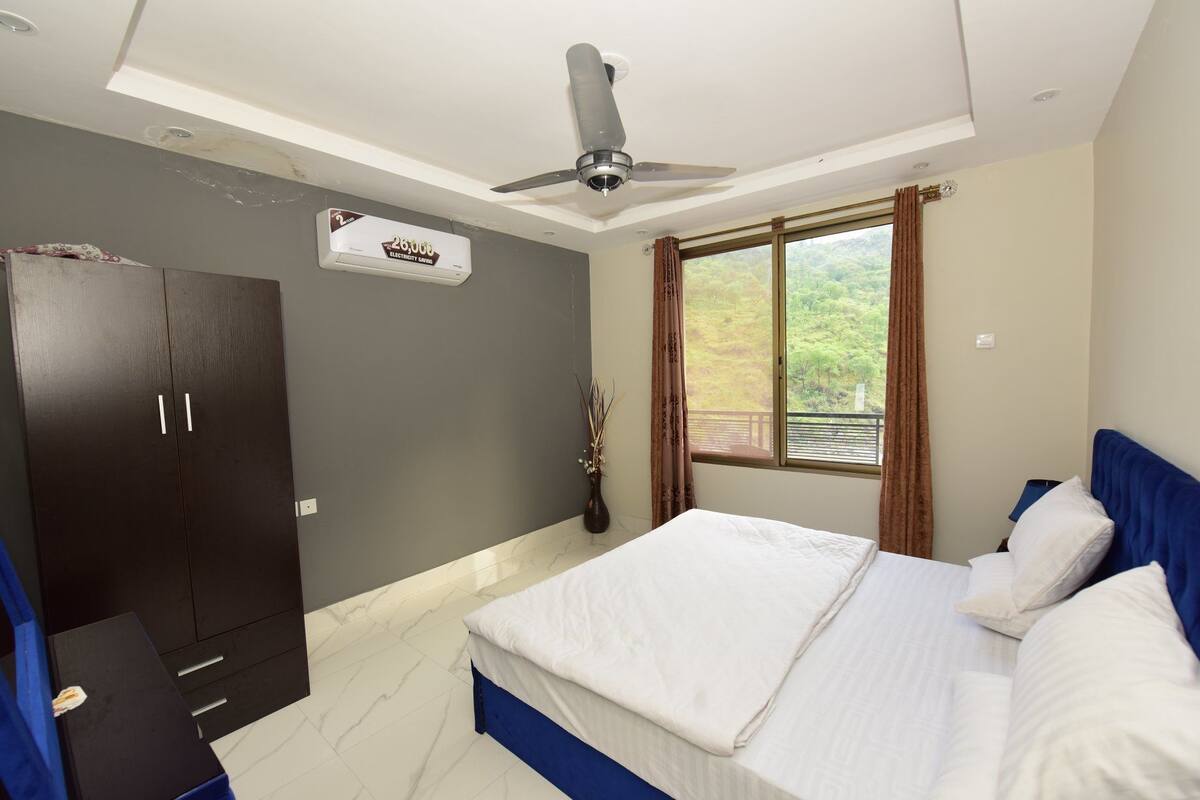 Une chambre à coucher comprenant un lit, une armoire, un ventilateur de plafond, une fenêtre avec des rideaux et une vue sur la verdure à l’extérieur.