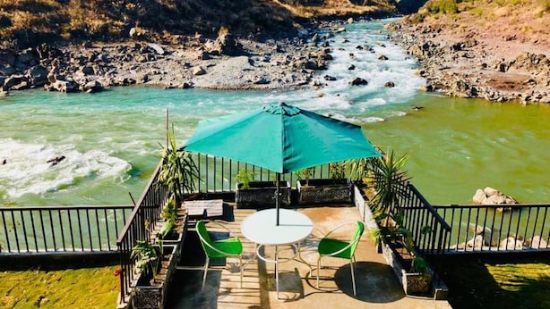 A terrace with a table and chairs, overlooking a river with a rocky riverbed and a bridge. - La Orilla Hotel (Muzaffarabad)