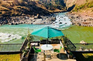 A terrace with a table and chairs, overlooking a river with a rocky riverbed and a bridge. - La Orilla Hotel (Muzaffarabad)