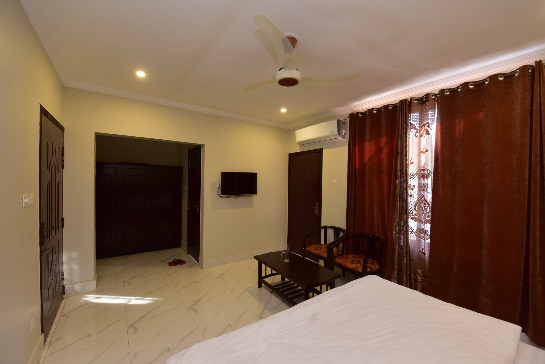 A hotel room with a bed, a desk with two chairs, a ceiling fan, and an air conditioning unit.
