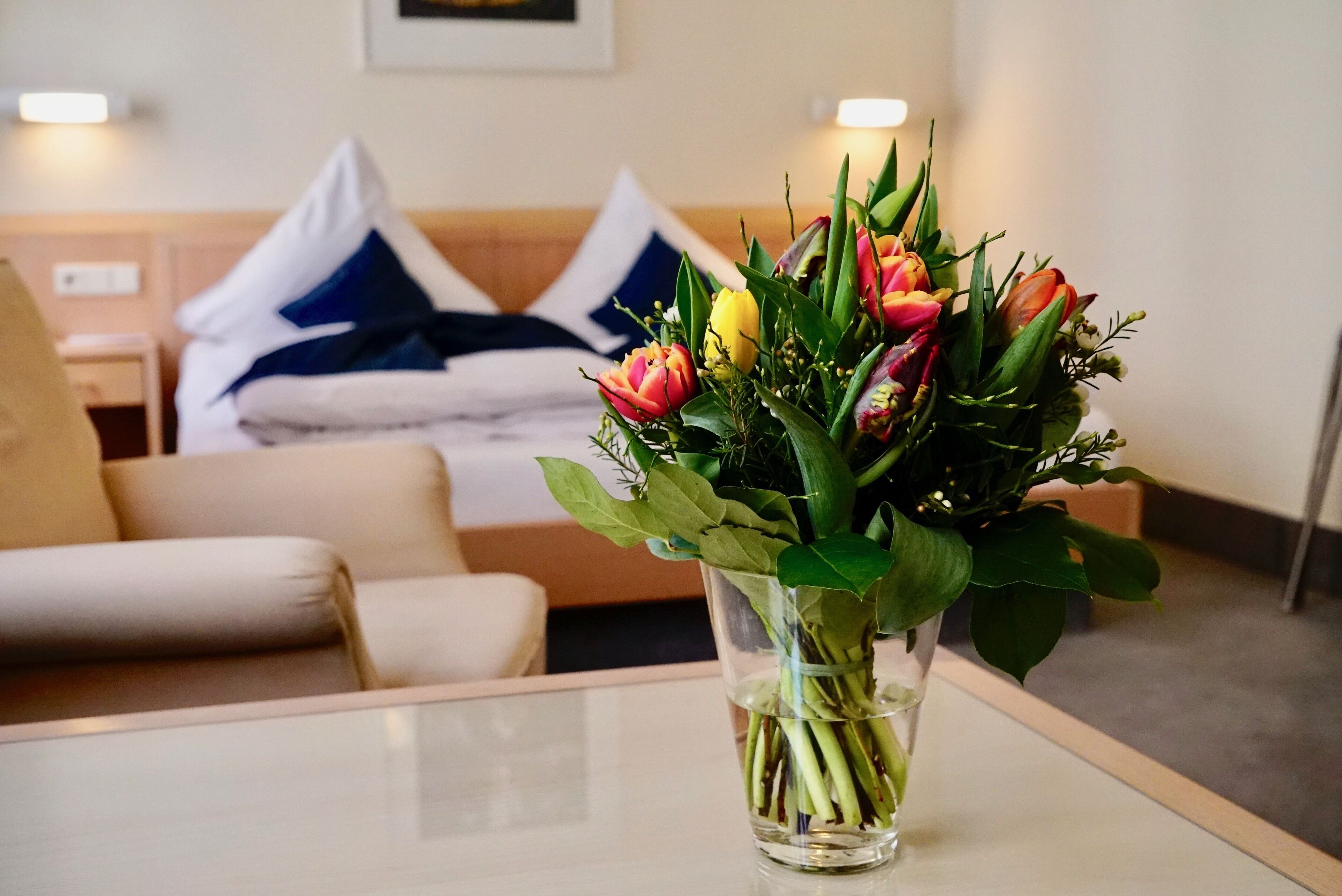 A hotel room with a bed, a bedside table with a vase of flowers, and a couch.