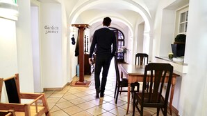 Un hombre caminando por un pasillo con piso de baldosas, sillas de madera y una mesa.