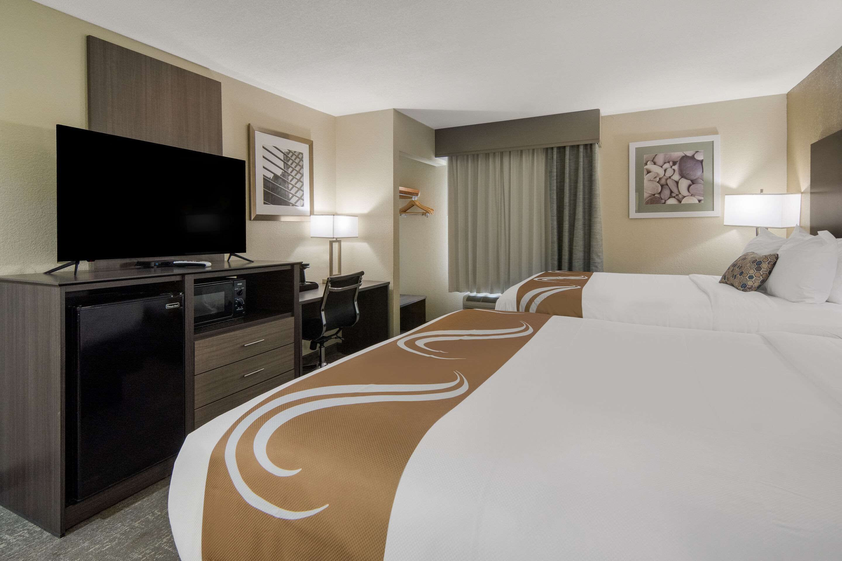 A hotel room with a large bed, a desk with a flat-screen TV, a mini-fridge, a chair, and a desk lamp.