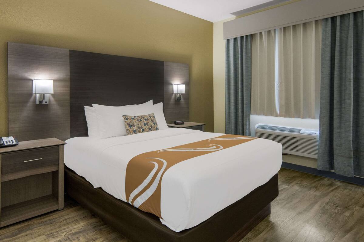 A hotel room with a large bed, two bedside lamps, a nightstand, a window with curtains, and an air conditioning unit.