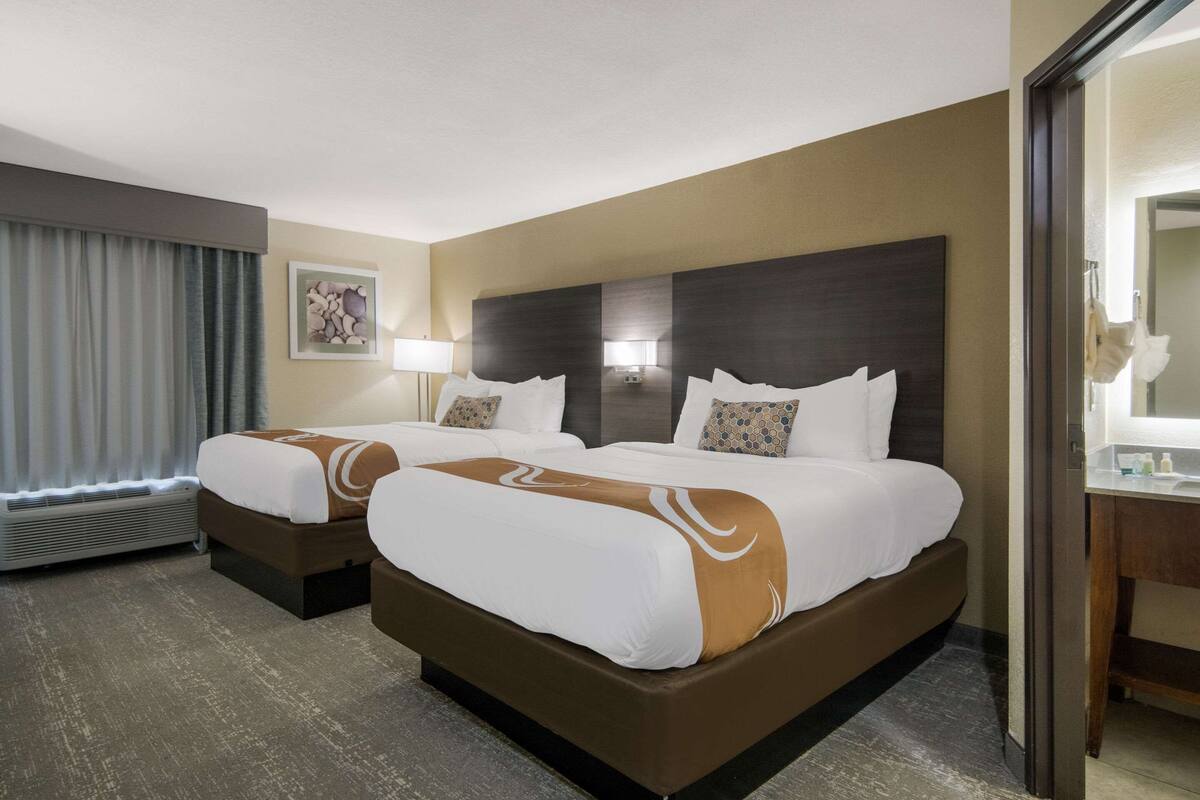 A hotel room with two beds, a large mirror, and a bathroom with a sink and mirror.
